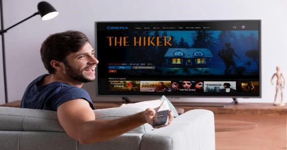 JalshaMoviezHD: Your Gateway to the World of High-Definition Entertainment 3 A person sitting on a couch facing a large TV screen displaying a movie selection interface with the title ‘THE HIKER’ highlighted. The screen also shows options like ‘Watch Now’ and ‘Already Purchased? Redeem Here.’ The individual is holding a tablet or remote control in a minimalistic living room.